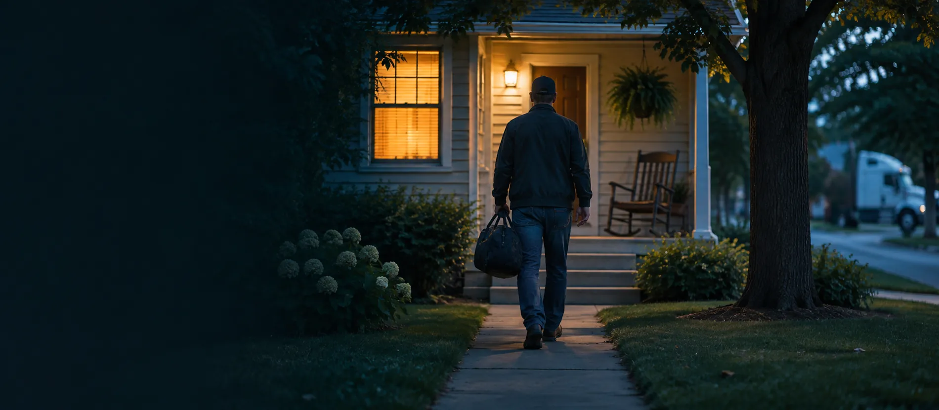 A driver with a duffel bag walks up to a lit front porch at dusk